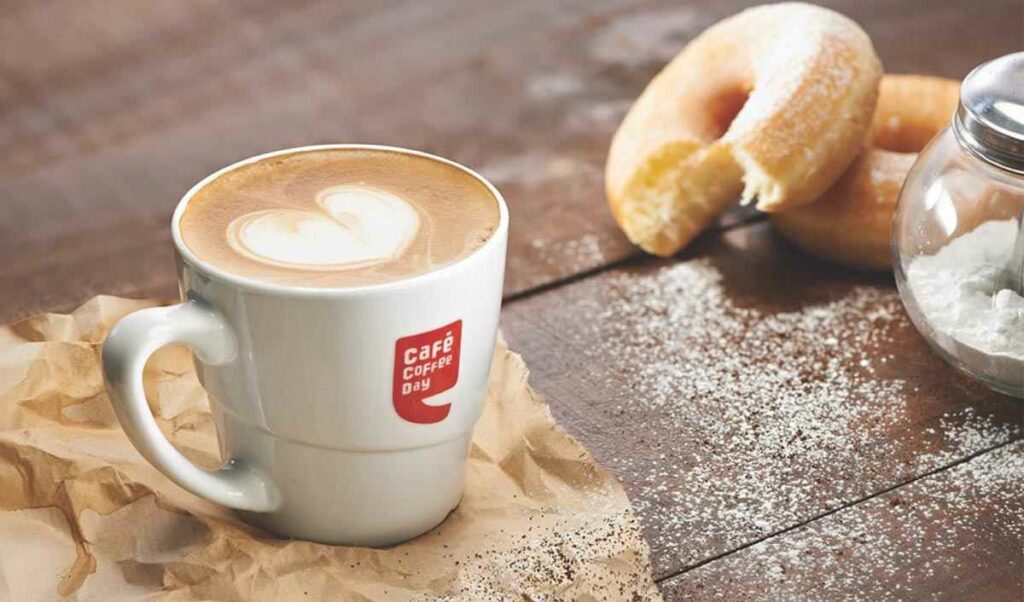 This image shows a cup of coffee with doughnuts in the background