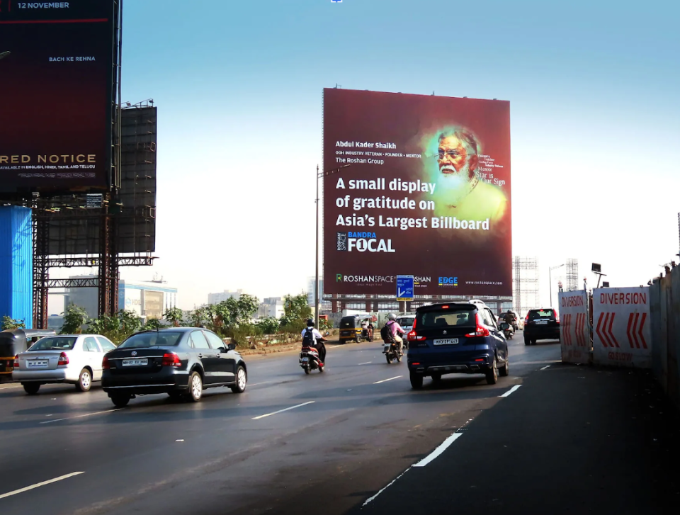 Large roadside billboard with a portrait and text, viewed from a busy multi-lane road with cars and motorcycles