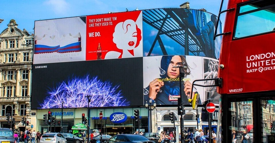 Large digital billboards displaying multiple brand advertisements at a busy city intersection with traffic and pedestrians visible