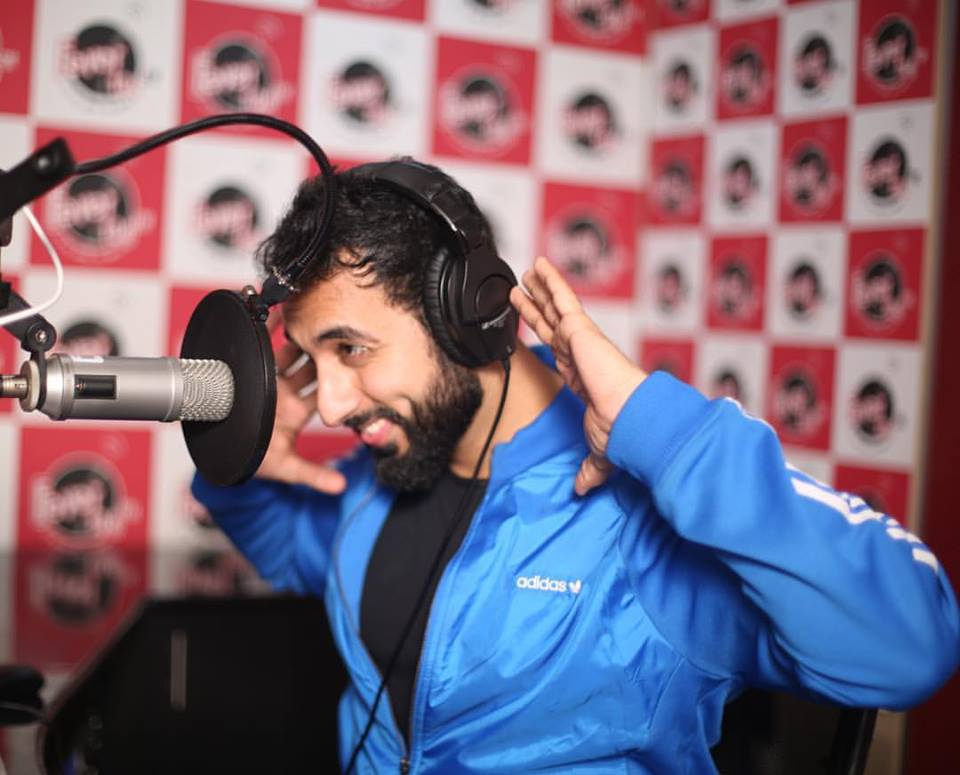 Radio Studio Advertising and Broadcast Radio host wearing headphones speaking into a microphone inside a recording studio with branded backdrop.