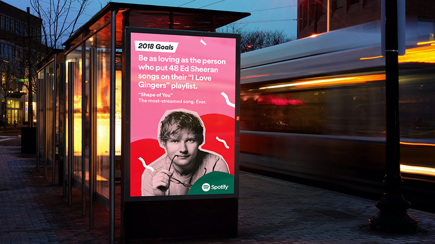 Spotify 2018 Goals Bus Shelter Advertisement Featuring Ed Sheeran Illuminated bus shelter advertisement featuring Spotify’s 2018 Goals campaign highlighting Ed Sheeran’s “Shape of You,” displayed on a digital panel at night with a passing bus in motion blur.