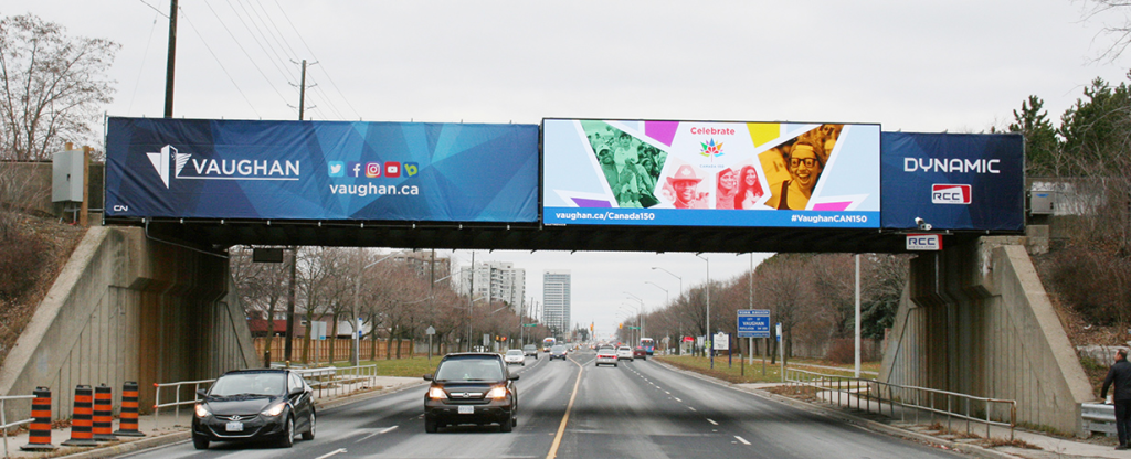 Bridge Billboard Advertising Vaughan Canada 150 Campaign Large bridge-mounted digital billboard spanning a multi-lane roadway displaying Vaughan city branding and a Canada 150 celebration campaign above passing traffic.