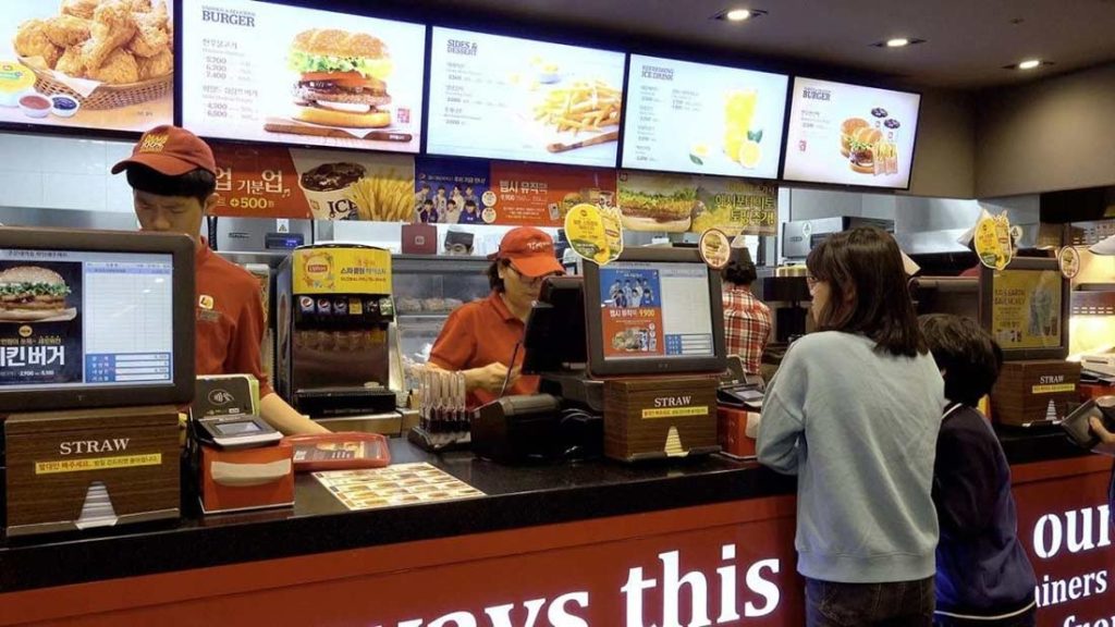 Quick Service Restaurant Ordering Counter Customers ordering food at a busy fast-food restaurant counter with digital menu boards displaying burgers, fries, and meal options.