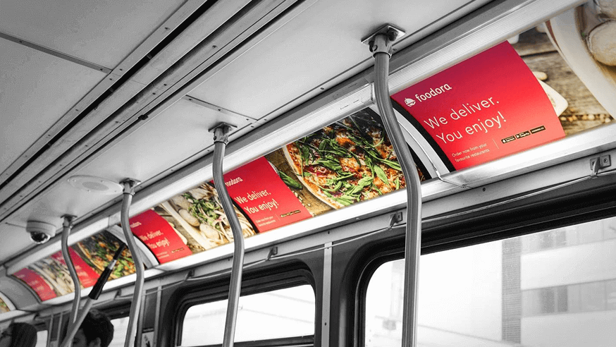 Foodora In-Transit Advertising Inside Public Bus Foodora food delivery advertisement displayed inside a city bus with red panels showing dishes and the message “We deliver. You enjoy.”