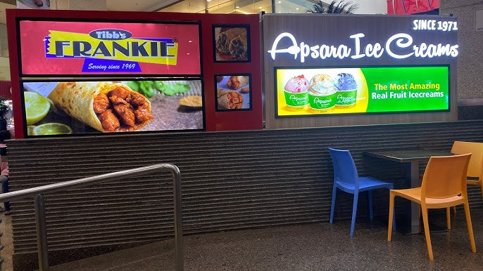 Bright digital advertising displays for Tibbs Frankie and Apsara Ice Creams at a food court seating area. Bright digital advertising displays for Tibbs Frankie and Apsara Ice Creams at a food court seating area.