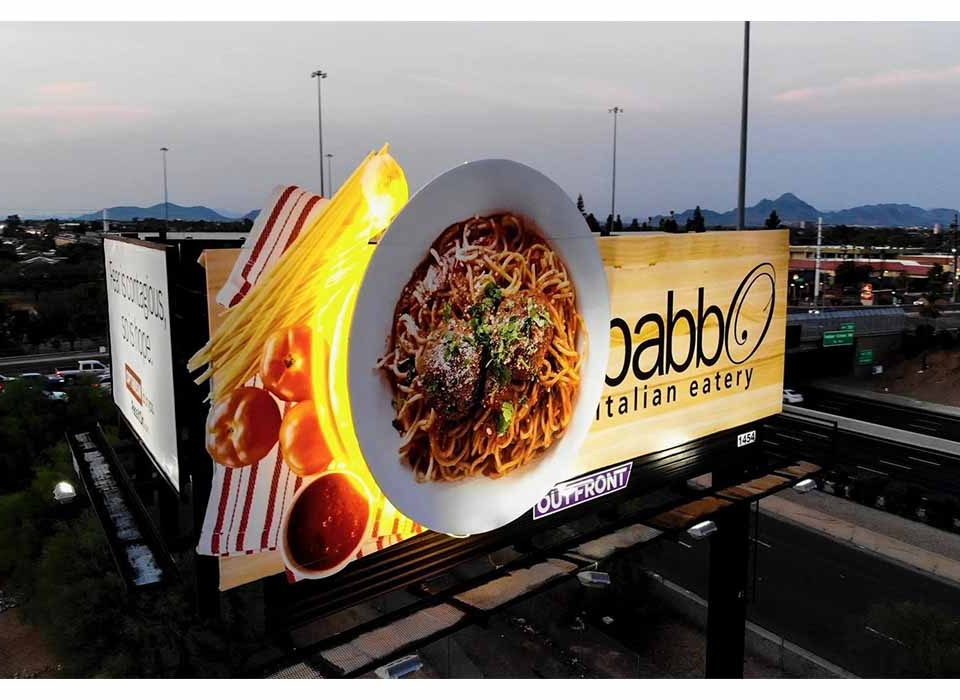 3D highway billboard advertising an Italian restaurant featuring an oversized plate of spaghetti with meatballs and illuminated pasta elements extending beyond the billboard.