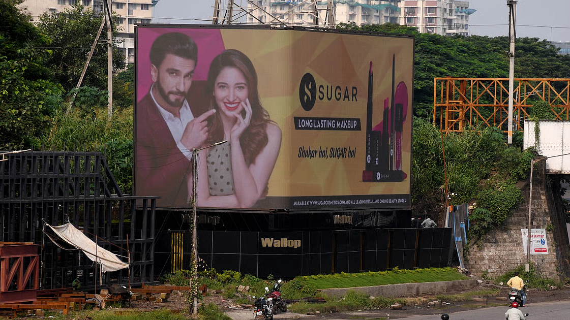 SUGAR Cosmetics Highway Billboard Display SUGAR Cosmetics billboard showcasing beauty products and brand ambassadors mounted on a roadside structure surrounded by greenery and urban buildings.