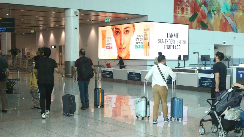Lakmé Sunscreen Digital Billboard at Airport Check-in Area Travelers with luggage walking through an airport terminal past a large Lakmé Sun Expert SPF 50 digital advertisement near the check-in counters.