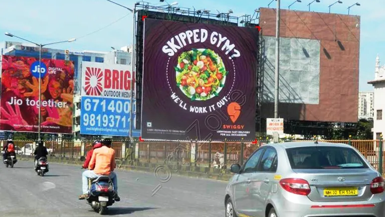 Swiggy Creative Outdoor Billboard Campaign Swiggy billboard reading “Skipped Gym? We’ll Work It Out” displayed along a busy road with cars and motorcycles passing underneath.