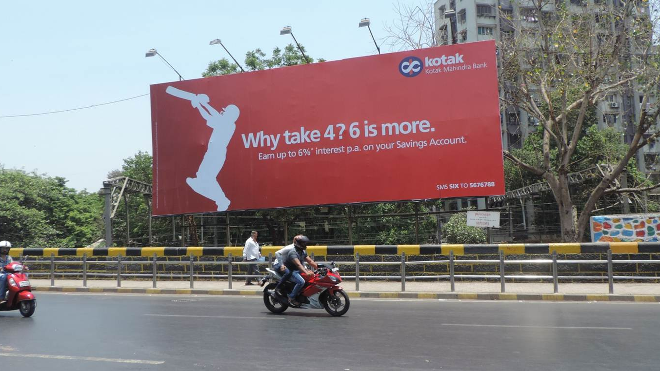 Kotak Mahindra Bank Financial Services Billboard Kotak Mahindra Bank billboard promoting savings account interest rates placed above a city roadway with motorcyclists riding below.