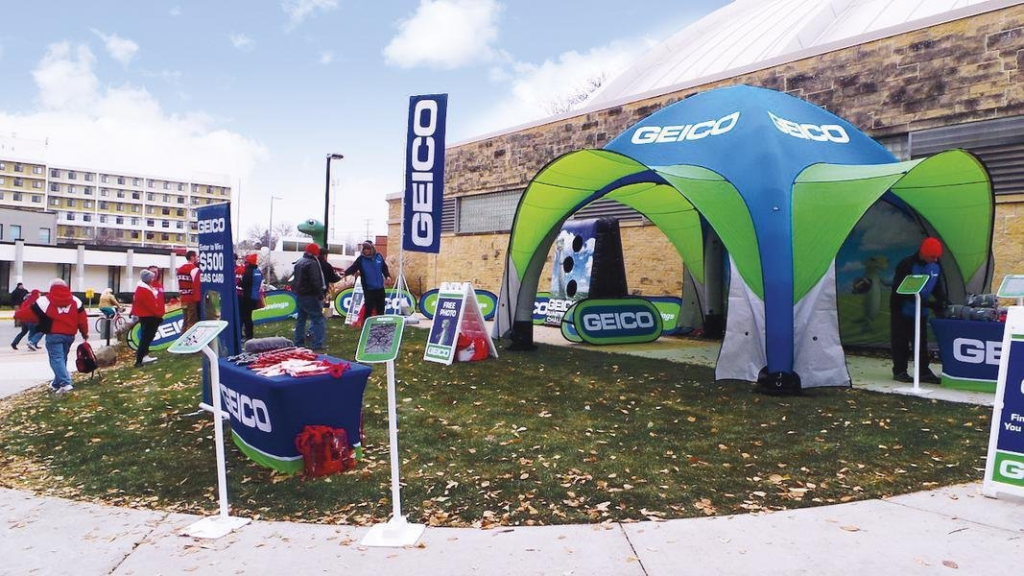 GEICO outdoor promotional booth setup for experiential marketing event. GEICO branded outdoor activation booth with promotional tent, signage, and staff engaging visitors during a marketing event.