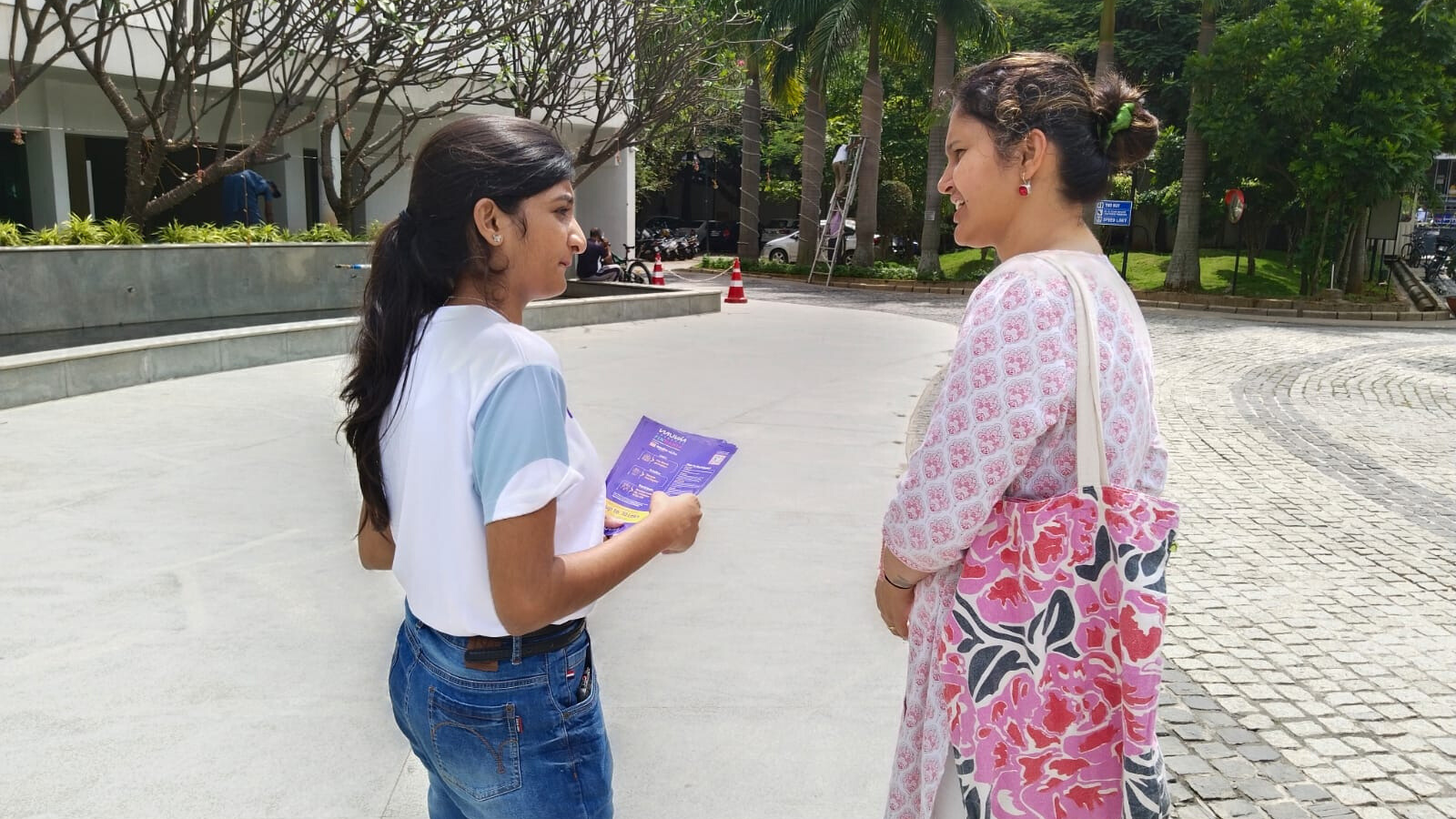 On-Ground Flyer Distribution Marketing Activity Young woman handing a promotional flyer to another woman in a residential outdoor setting as part of direct marketing activity.