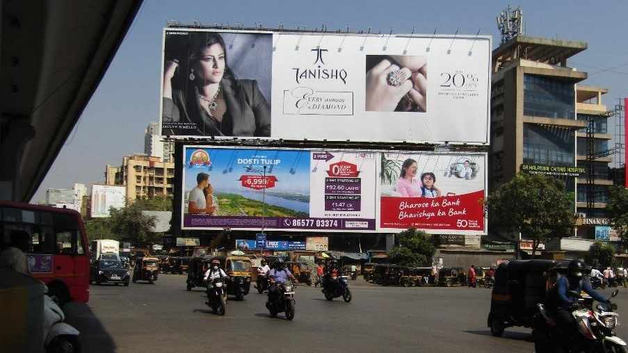 Tanishq Jewellery Billboard at City Intersection Large Tanishq jewellery billboard mounted above a busy city intersection with multiple vehicles, auto-rickshaws, and surrounding commercial buildings.